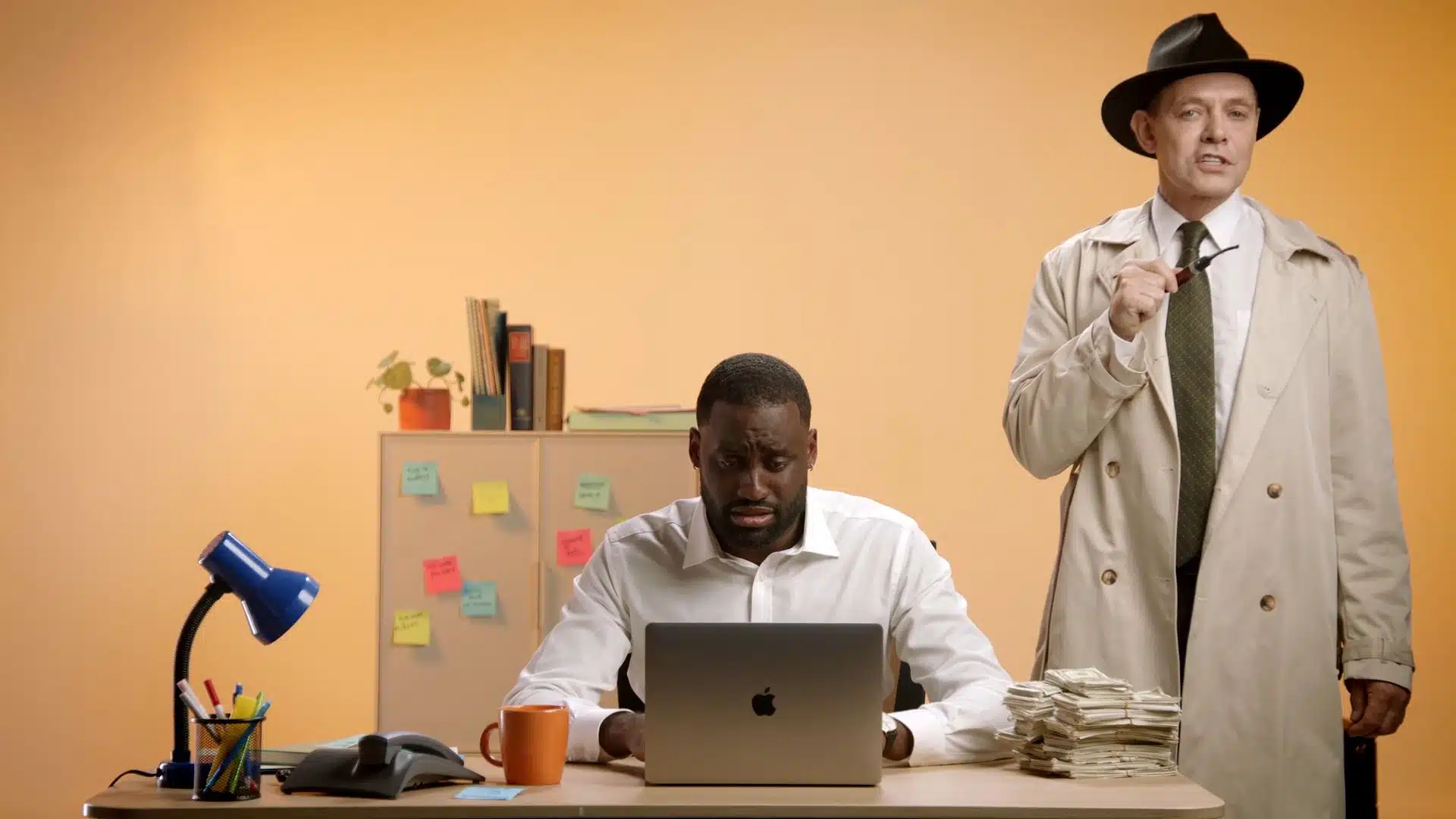 Man infront of a laptop on a desk, while another man in a trench coat holding a pipe