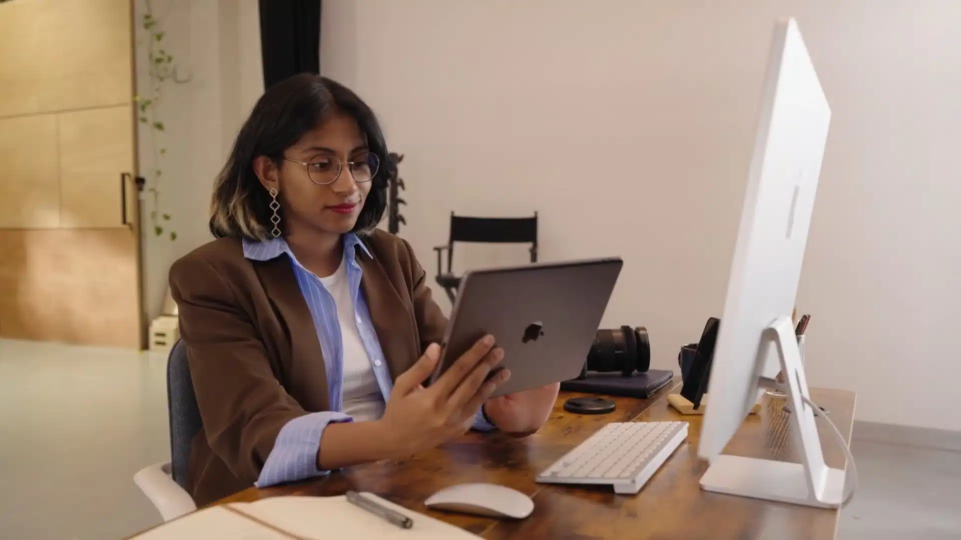 Woman in glasses and a blazer working at a desk, holding an iPad