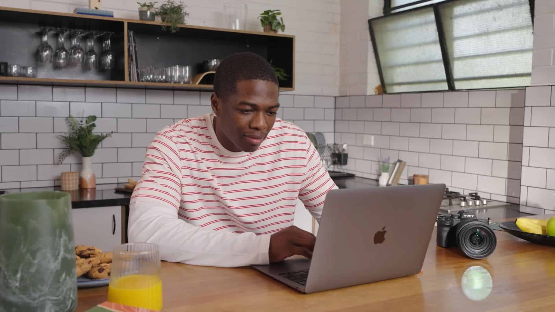 Man sitting in kitchen working on laptop with camera