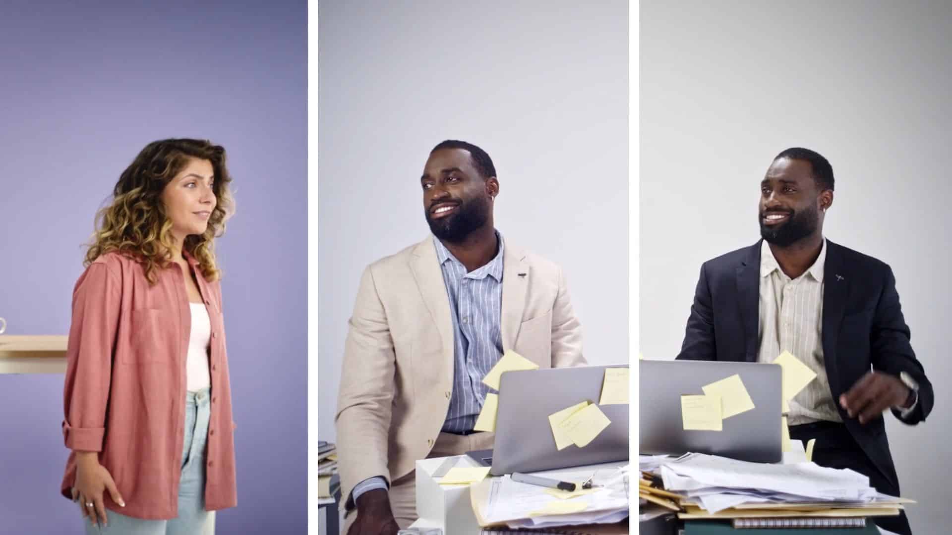 woman standing and smiling at two men sitting at cluttered desks with laptops