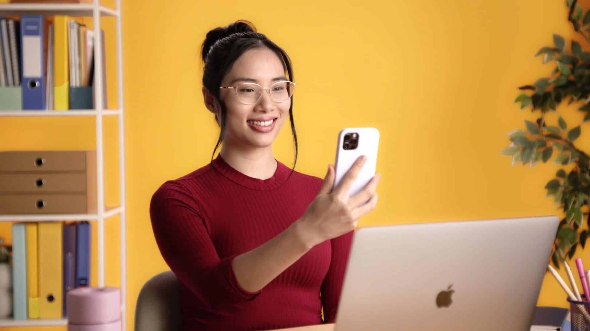 Woman smiling while using smartphone at desk with laptop in a bright home office