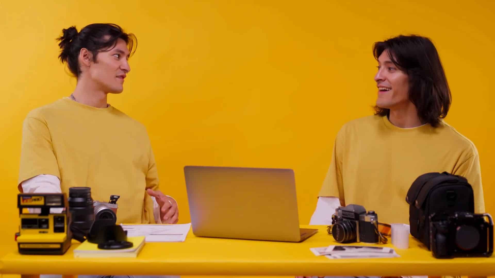 Two people in yellow shirts discussing cameras and a laptop at a yellow table